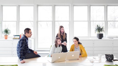 A group of young professionals gather around two laptops in an office with white storage units on the walls and large windows to the outside.
