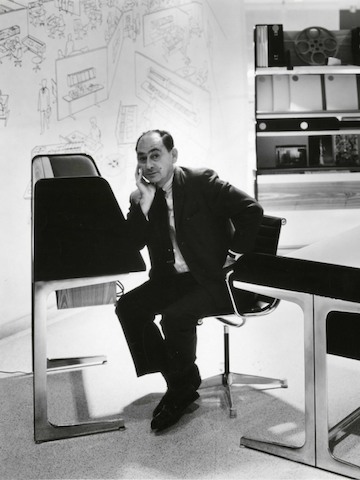 Designer George Nelson leans on an Action Office standing desk with sketches of workplace settings and furniture on the wall behind him.