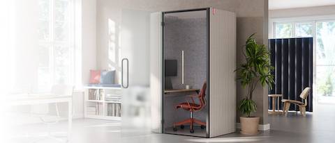 An open office, featuring an enclosed Bay Work Pod with red task chair pulled up to a work surface with computer monitor.