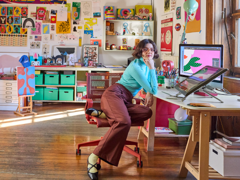 Olimpia Zagnoli arbeitet in ihrem großen, sonnendurchfluteten Atelier an einem Computer vor einem Fenster, umgeben von bunten Kunstwerken und Objekten.