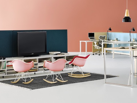 Three pink Eames Molded Plastic rocking chairs facing a teleconferencing monitor that is supported by a Canvas Office Landscape storage unit.