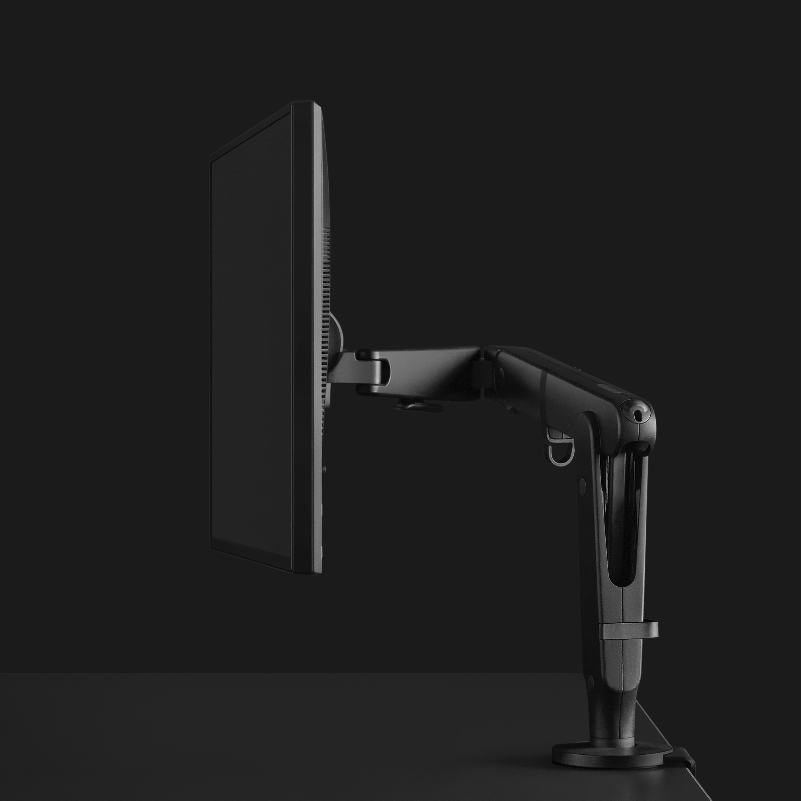 Dark grey Ollin Monitor Arm shown from the side with a dramatically lit black background.
