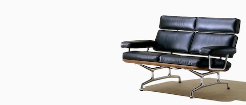 Angled view of a black leather Eames loveseat with polished aluminum legs and arm supports.