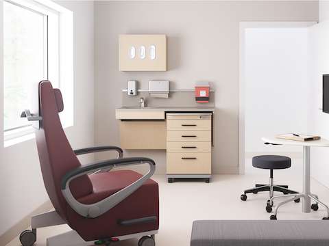 An exam room setting featuring Mora casework in an ash wood finish with a supply cart, Intent Solution in an ash finish, a physician's stool in dark gray, an Ava recliner in a maroon upholstery, and a Riva bench in gray.