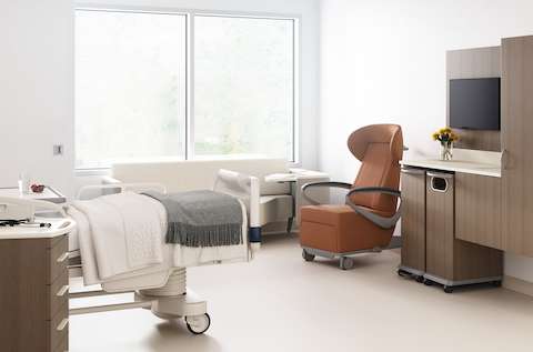 A patient room with a footwall of Mora casework with a trash cart and linen cart in medium brown wood finish, an Ava recliner in tan, a Palisade Flop Sofa in off white, a patient bed, with a Mora supply cart next to it.