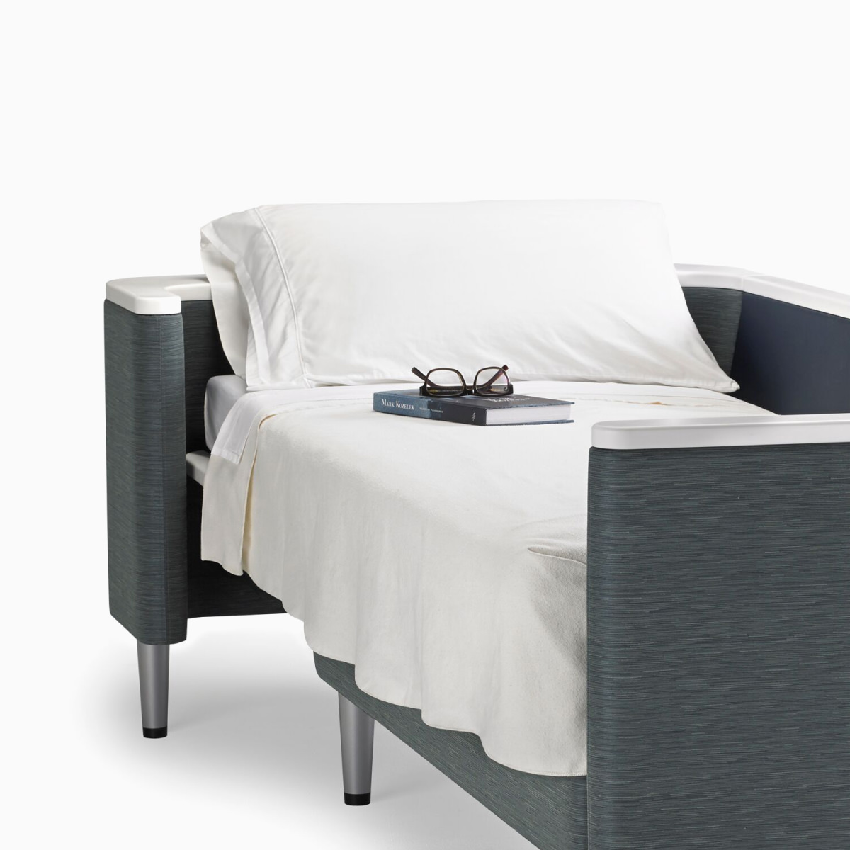 A Nemschoff Palisade Flop sofa converted to sleep surface in dark gray textile with white solid surface arms caps and white pillow and sheet.