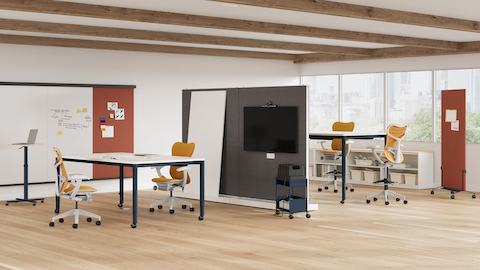 Open plan agile team space shown with two 4-leg height adjustable meeting tables featuring white surfaces, dark blue legs and technology buckets in center of tables. Room is split by sub-architectural boundary that features a monitor for video conferencing.