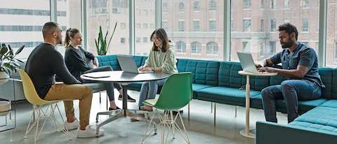 Three people are chatting in a casual lounge area in office while another person works on a laptop nearby. Three people sit on a turquoise Tuxedo Sofa while one person sits on a yellow Eames Shell Chair. 