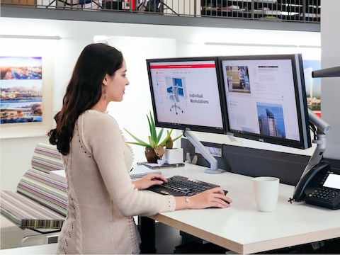 Woman standing at a sit-to-stand desk, looking at two computer monitors. 