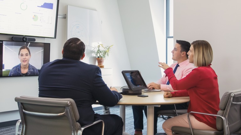 Three people sitting in gray Eames Soft Pad Chairs talking with a remote colleague whose face appears on a large monitor on the wall.