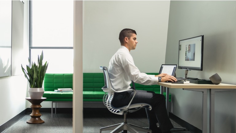 Man in a private office working on a laptop at an Everywhere Table and sitting in a gray Setu Chair with a green Tuxedo Sofa in the background.