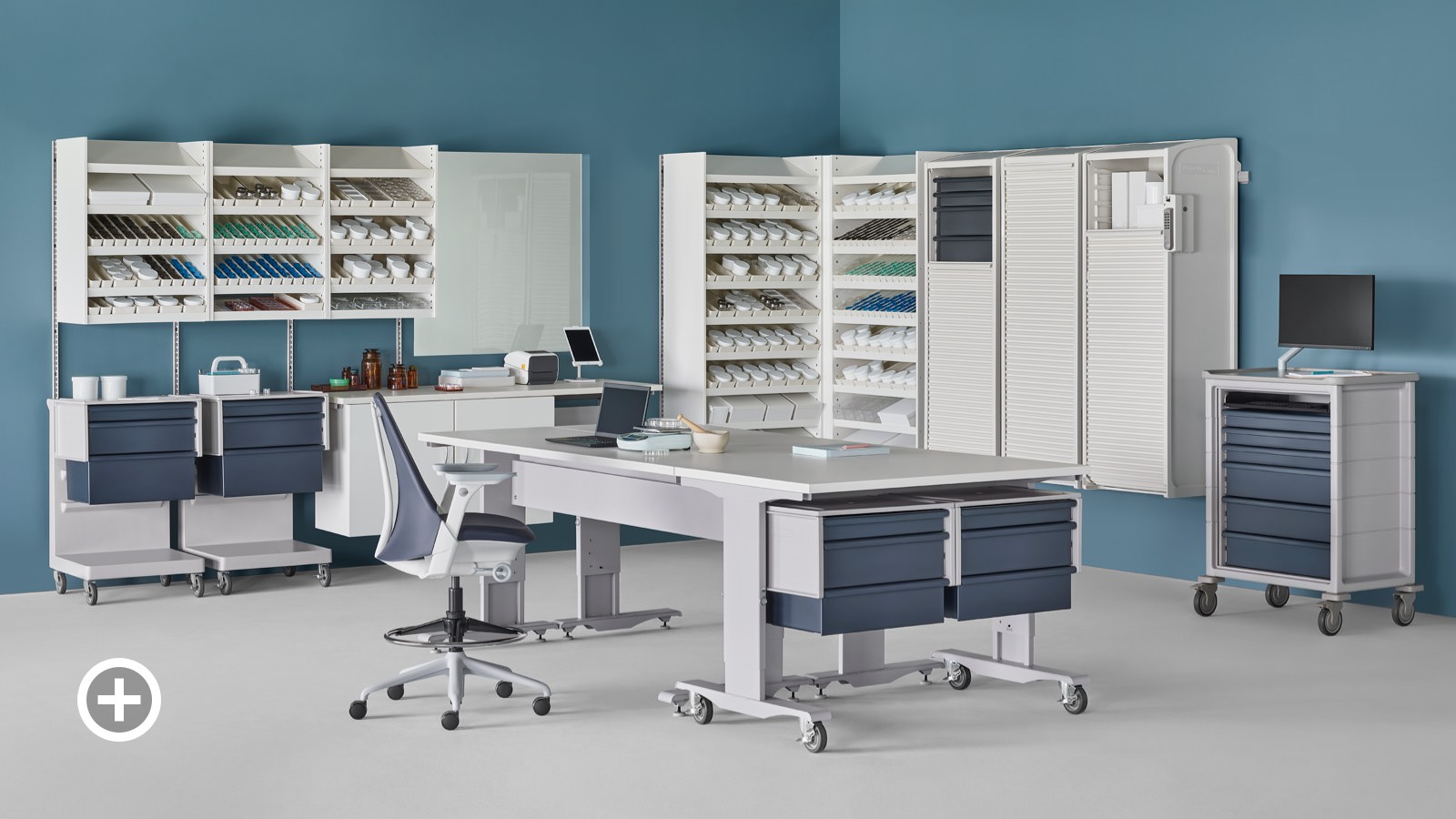 A pharmacy containing a white Co/Struc System of dispensing shelves, lockers, process tables, L Carts with blue drawers, a technology cart with blue drawers and a Sayl Stool.