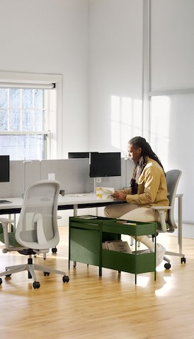 A person sits at a grey OE1 Rectangular Table joined with other tables to form a bench, with a grey OE1 Project Table and green Lino Chairs in the foreground and an OE1 Wall Rail and Board in the background.