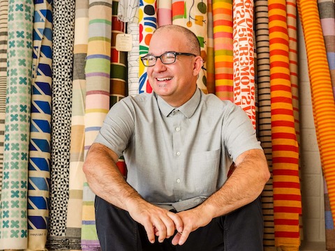 Vince Bravo sits in front of large rolls of colorful patterned textiles by Alexander Girard.