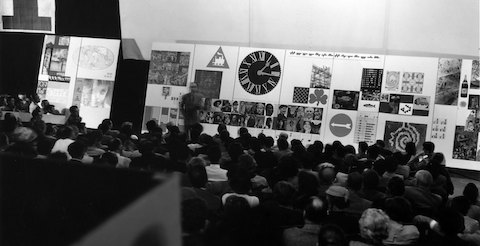 A black-and-white image of a man speaking before a seated crowd.