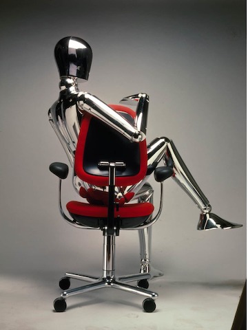 An archival studio photo of a red Ergon Chair from the back, with a silver mannequin sitting slouched in the chair.