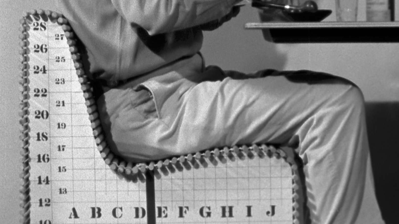 A close-up archival black and white photo of a man sitting in an ergonomic measuring device pulled up to a dinner table.