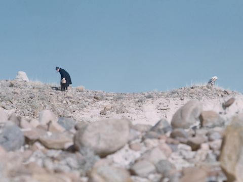 Ray Eames bending over in a field of rocks.