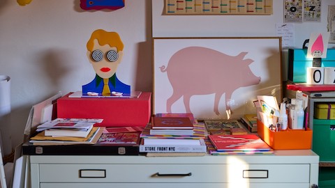 Stacks of books, notes, artist supplies and colorful art on top of a gray flat-file cabinet.