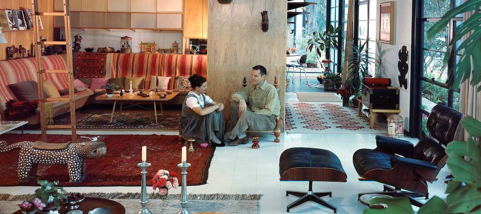 In this color photo, Charles and Ray Eames sit on the floor in their living room. They are surrounded by an Eames Lounge Chair and Ottoman and objects from their travels.