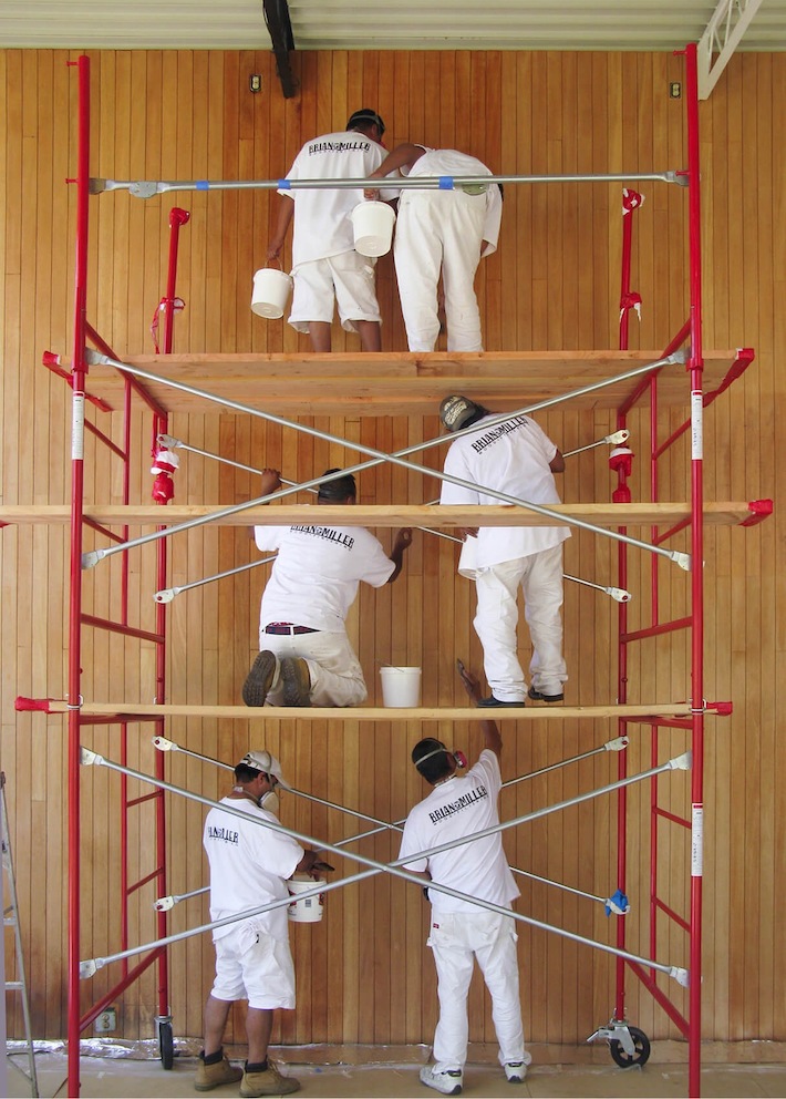A color photo of six men on a red scaffold working on repairs to the wall in the living room of the Eames House. 