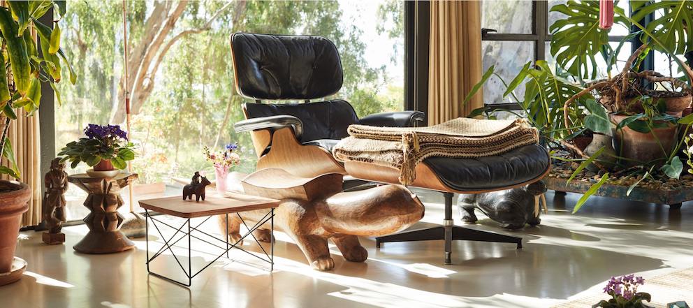 A color photo of a black leather Eames Lounge Chair and Ottoman in the living room at the Eames House. A tan blanket is folded on the ottoman, and a LTR table sits nearby. 