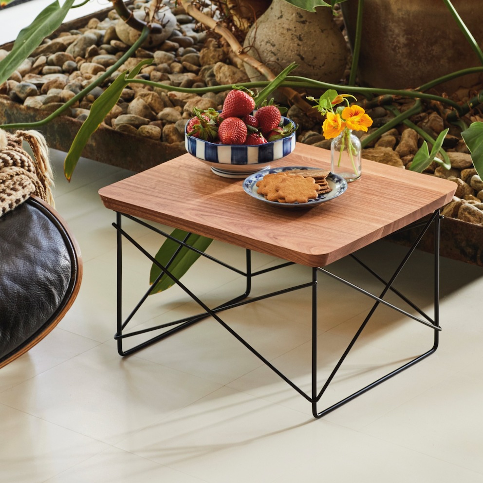 A close-up, color side-view photo of an Eames LTR Table topped with a vase of yellow flowers, a blue and white bowl of strawberries, and a blue and white plate of cookies. 